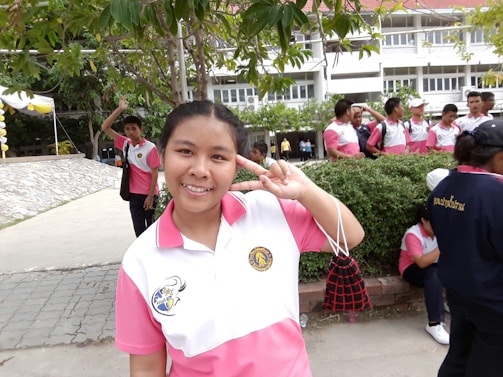 A group of people wearing white and pink uniforms are gathered outdoors in what appears to be a school setting. A person in the foreground is smiling and making a peace sign. The background includes more people, trees, and a building with windows.