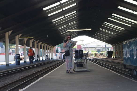 A person stands on a train platform holding a map, wearing sunglasses and casual summer attire. The platform is covered with a large roof, and railway tracks run parallel to it. There are a few people scattered in the background, including someone in an orange vest. The environment is well-lit with natural light coming through the roof sections.