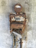 An old, rusted industrial machine mounted on a concrete wall with visible dials and exposed internal components. The metal surface shows significant corrosion and decay, indicating many years of weathering.