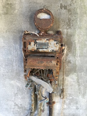 An old, rusted industrial machine mounted on a concrete wall with visible dials and exposed internal components. The metal surface shows significant corrosion and decay, indicating many years of weathering.