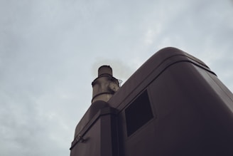 A smokestack on an industrial building emits faint smoke against a cloudy sky backdrop. The structure appears metallic with rivets and a rounded edge design.