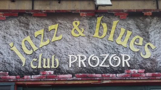 A sign features the words 'jazz & blues club PROZOR' in large, stylized letters. The background resembles a brick wall with some painted areas.