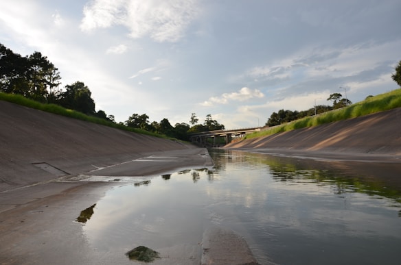 A wide concrete channel with gentle slopes on either side, filled partially with water that reflects the sky. Surrounding the channel are lush green grass and trees, with a bridge crossing in the background. The sky is partly cloudy, creating a serene atmosphere.