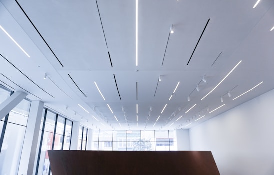 A modern interior space with a high ceiling featuring linear LED lighting strips and spotlights. The room has large floor-to-ceiling windows letting in natural light. The architecture is minimalist with an emphasis on clean, geometric lines.