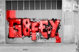 Large graffiti with the word 'GUEFY' in vibrant red, covering a ribbed metal garage door. The graffiti includes abstract shapes and tags, surrounded by grayscale elements such as the sidewalk and adjacent wall. A slightly defocused street scene is seen in black and white, with buildings and parked vehicles visible.