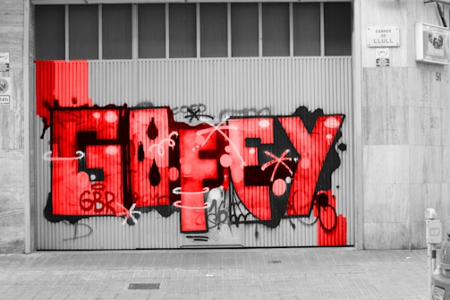 Large graffiti with the word 'GUEFY' in vibrant red, covering a ribbed metal garage door. The graffiti includes abstract shapes and tags, surrounded by grayscale elements such as the sidewalk and adjacent wall. A slightly defocused street scene is seen in black and white, with buildings and parked vehicles visible.