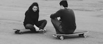 Two individuals are seated on skateboards on a paved surface. One person appears to be injured, holding their ankle and displaying a pained expression, while the other sits beside them with their back turned.