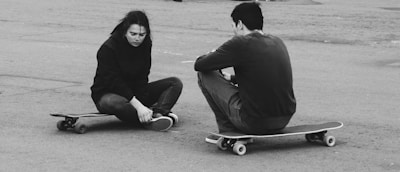 Two individuals are seated on skateboards on a paved surface. One person appears to be injured, holding their ankle and displaying a pained expression, while the other sits beside them with their back turned.