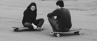 Two individuals are seated on skateboards on a paved surface. One person appears to be injured, holding their ankle and displaying a pained expression, while the other sits beside them with their back turned.