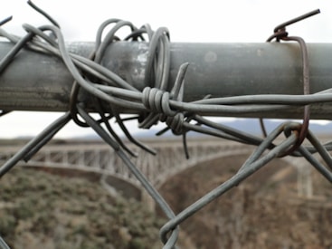 A section of a wire fence is secured with thick metal wire, creating a close-up view that highlights the intertwined and rusted loops. In the blurred background, a bridge spans across a canyon, cutting across a rugged landscape.