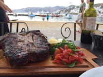 A large, grilled steak is served on a wooden board accompanied by a fresh salad of arugula and cherry tomatoes. Parmesan shavings are placed on the side. In the background, a scenic beach setting with boats on calm water is visible. A bottle of red wine and a basket of bread are also present on the table.
