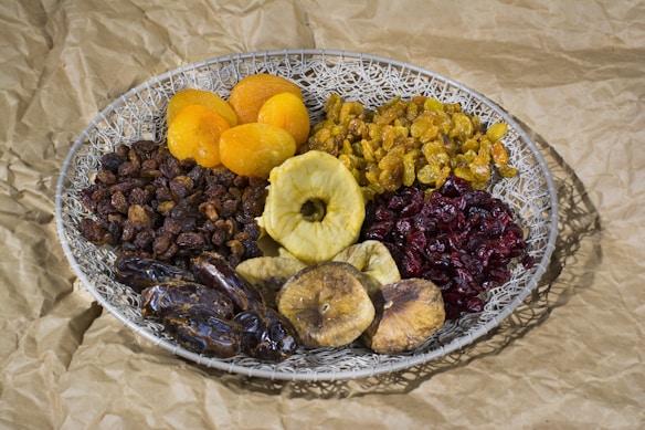 A woven basket filled with an assortment of dried fruits including apricots, golden raisins, cranberries, dates, figs, and a slice of dried apple. The basket is placed on crumpled brown paper that gives a rustic look.
