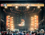 A vibrant outdoor music festival stage with large screens displaying graphics and lighting fixtures set up above. A crowd of people is gathered at the front, creating a lively and energetic atmosphere.