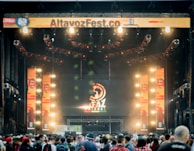 A vibrant outdoor music festival stage with large screens displaying graphics and lighting fixtures set up above. A crowd of people is gathered at the front, creating a lively and energetic atmosphere.