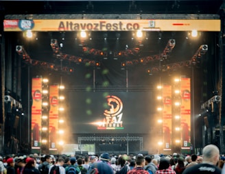 A vibrant outdoor music festival stage with large screens displaying graphics and lighting fixtures set up above. A crowd of people is gathered at the front, creating a lively and energetic atmosphere.