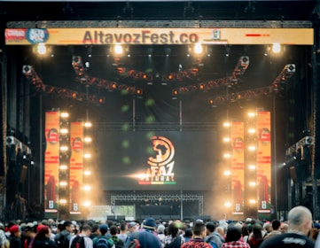 A vibrant outdoor music festival stage with large screens displaying graphics and lighting fixtures set up above. A crowd of people is gathered at the front, creating a lively and energetic atmosphere.