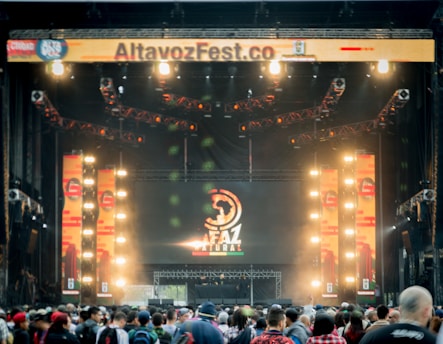 A vibrant outdoor music festival stage with large screens displaying graphics and lighting fixtures set up above. A crowd of people is gathered at the front, creating a lively and energetic atmosphere.