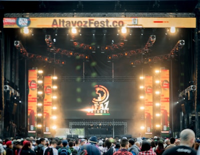 A vibrant outdoor music festival stage with large screens displaying graphics and lighting fixtures set up above. A crowd of people is gathered at the front, creating a lively and energetic atmosphere.