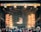 A vibrant outdoor music festival stage with large screens displaying graphics and lighting fixtures set up above. A crowd of people is gathered at the front, creating a lively and energetic atmosphere.