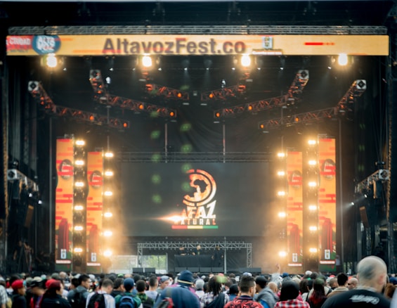 A vibrant outdoor music festival stage with large screens displaying graphics and lighting fixtures set up above. A crowd of people is gathered at the front, creating a lively and energetic atmosphere.