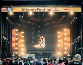 A vibrant outdoor music festival stage with large screens displaying graphics and lighting fixtures set up above. A crowd of people is gathered at the front, creating a lively and energetic atmosphere.