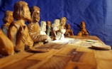 A detailed wooden sculpture featuring multiple figures seated around a long table, resembling a famous biblical scene. The intricate carvings capture expressions and details of each figure, and the craftsmanship highlights the wood grain and carving techniques.