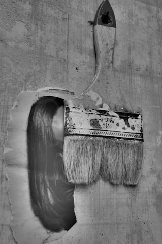 An artistic image of an old, used paintbrush seemingly brushing across an old paper or wall surface. Interestingly, part of a woman's face with long hair is seamlessly integrated with the brush stroke, creating a surreal visual effect. The image is in black and white, adding a nostalgic and mysterious atmosphere.