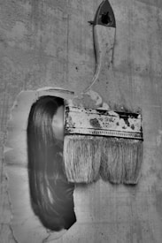 An artistic image of an old, used paintbrush seemingly brushing across an old paper or wall surface. Interestingly, part of a woman's face with long hair is seamlessly integrated with the brush stroke, creating a surreal visual effect. The image is in black and white, adding a nostalgic and mysterious atmosphere.