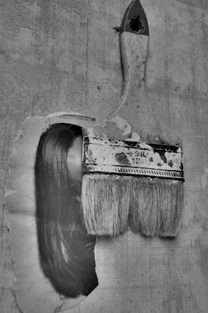 An artistic image of an old, used paintbrush seemingly brushing across an old paper or wall surface. Interestingly, part of a woman's face with long hair is seamlessly integrated with the brush stroke, creating a surreal visual effect. The image is in black and white, adding a nostalgic and mysterious atmosphere.
