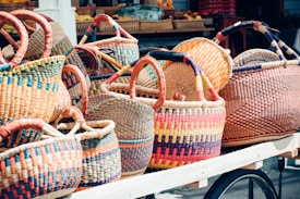 A variety of colorful woven baskets with intricate patterns and designs are displayed on a wooden cart. The baskets feature a rich mix of colors including orange, red, blue, and yellow, and have sturdy-looking handles. The background includes more baskets and some fruits, suggesting a market or outdoor setting.
