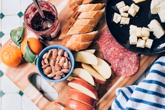 A wooden cutting board is filled with a variety of snacks. Sliced baguette bread, apple slices, a blue bowl of almonds, and two tangerines are arranged artistically. A jar of red jam with a spoon inside sits next to the fruit. Salami slices and cubes of cheese are placed on a black plate. A striped cloth is partially visible on the side.