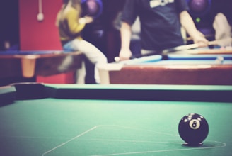 A billiard table with an 8-ball in focus, while two people are visible in the background, blurred to show motion or activity. The scene conveys a casual atmosphere, possibly in a game room or recreational facility.