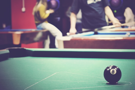 A billiard table with an 8-ball in focus, while two people are visible in the background, blurred to show motion or activity. The scene conveys a casual atmosphere, possibly in a game room or recreational facility.