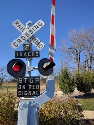 A railway crossing sign stands prominently, featuring a pair of round, red warning lights positioned on either side. Below the cross-shaped 'RAILROAD CROSSING' sign, there are indications for '2 TRACKS' and an instruction to 'STOP ON RED SIGNAL'. The background includes leafless trees and bushes under a clear blue sky.