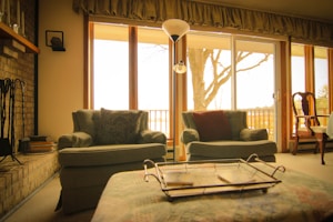 A cozy living room with vintage armchairs positioned near large windows that allow natural light to flood the space. A fireplace with books and fire tools rests to the side, and a decorative tray sits on a cushioned ottoman. A tree outside is visible through the window, hinting at a serene outdoor setting. Elements of traditional decor, like a wooden chair with armrests, add to the homely atmosphere.