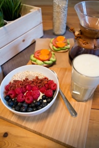 A breakfast scene featuring a bowl filled with yogurt, granola, raspberries, blueberries, and goji berries. Beside it, a glass of milk sits on a wooden board. In the background, there are avocado, tomato, and carrot open-faced sandwiches, a wooden crate with green plants, and a glass container of oats.