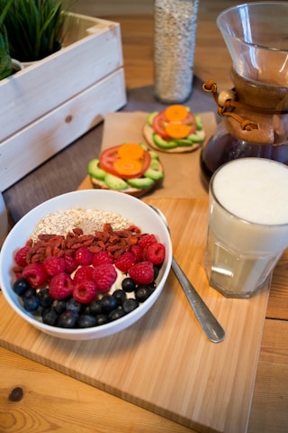 A breakfast scene featuring a bowl filled with yogurt, granola, raspberries, blueberries, and goji berries. Beside it, a glass of milk sits on a wooden board. In the background, there are avocado, tomato, and carrot open-faced sandwiches, a wooden crate with green plants, and a glass container of oats.
