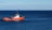 A bright red ship is navigating on a vast expanse of deep blue ocean. In the background, there is an oil rig visible on the horizon under a clear blue sky.