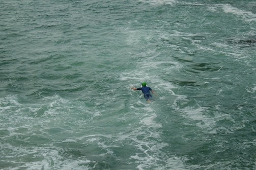 A person wearing a green helmet and dark wetsuit is swimming in a vast expanse of choppy, green ocean water.