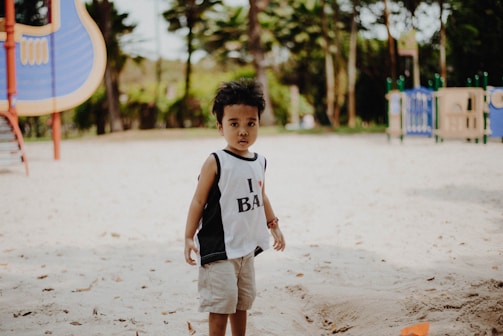 A young child stands in the foreground on a sandy playground. The child is wearing a sleeveless top and shorts. In the background, there are playground structures, including a slide and climbing equipment, set among trees and greenery under a clear sky.