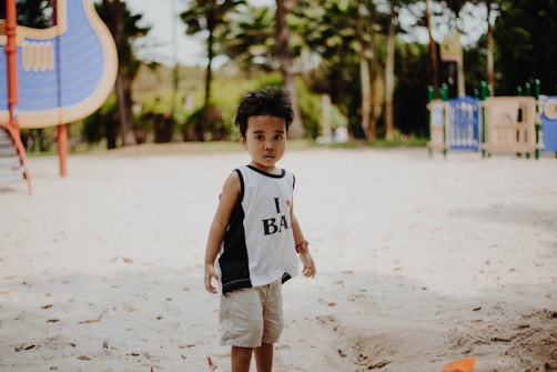 A young child stands in the foreground on a sandy playground. The child is wearing a sleeveless top and shorts. In the background, there are playground structures, including a slide and climbing equipment, set among trees and greenery under a clear sky.