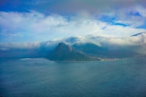 A majestic mountain rising above a calm sea, partially enveloped by thick, rolling clouds. The scene conveys a sense of tranquility and awe as the clouds create a dramatic backdrop to the landscape. The coastline is visible, with subtle details of a town or village at the base of the mountain, which adds a sense of scale and perspective.