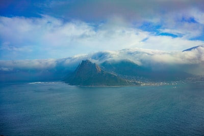 A majestic mountain rising above a calm sea, partially enveloped by thick, rolling clouds. The scene conveys a sense of tranquility and awe as the clouds create a dramatic backdrop to the landscape. The coastline is visible, with subtle details of a town or village at the base of the mountain, which adds a sense of scale and perspective.
