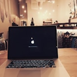 A dimly lit cafe interior with hanging light bulbs and modern decor features a central laptop on a wooden table with a visible Apple logo on the screen. In the background, there are plants, shelves with merchandise, and a person standing at the counter.