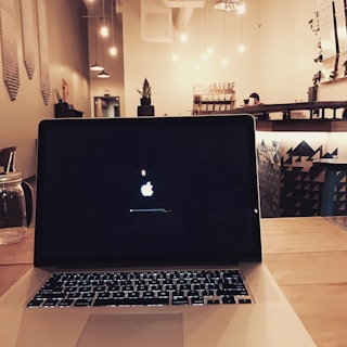 A dimly lit cafe interior with hanging light bulbs and modern decor features a central laptop on a wooden table with a visible Apple logo on the screen. In the background, there are plants, shelves with merchandise, and a person standing at the counter.