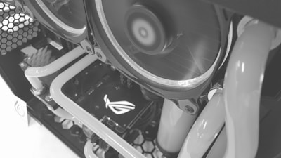 Close-up view of a computer's internal components featuring liquid cooling tubes and cooling fans. The ASUS Republic of Gamers (ROG) logo is visible on a hardware component, likely the graphics card. The image is in grayscale, highlighting the intricate design and layout of the PC internals.