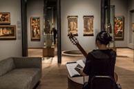 A person sits in an art gallery or museum, playing a guitar while facing a music stand with sheet music. The room has several paintings on the walls and sculptures inside glass cases. The lighting is focused on the artwork, highlighting the details and creating a cozy atmosphere.