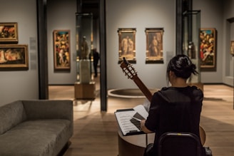A person sits in an art gallery or museum, playing a guitar while facing a music stand with sheet music. The room has several paintings on the walls and sculptures inside glass cases. The lighting is focused on the artwork, highlighting the details and creating a cozy atmosphere.
