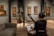 A person sits in an art gallery or museum, playing a guitar while facing a music stand with sheet music. The room has several paintings on the walls and sculptures inside glass cases. The lighting is focused on the artwork, highlighting the details and creating a cozy atmosphere.