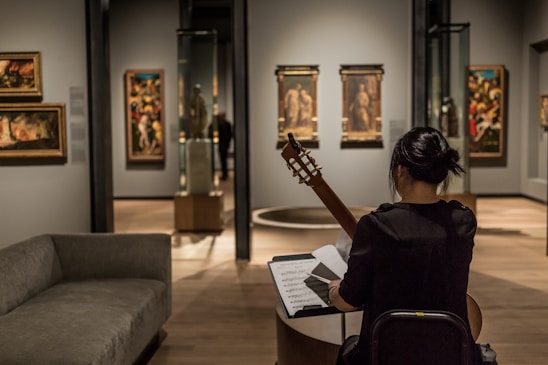 A person sits in an art gallery or museum, playing a guitar while facing a music stand with sheet music. The room has several paintings on the walls and sculptures inside glass cases. The lighting is focused on the artwork, highlighting the details and creating a cozy atmosphere.